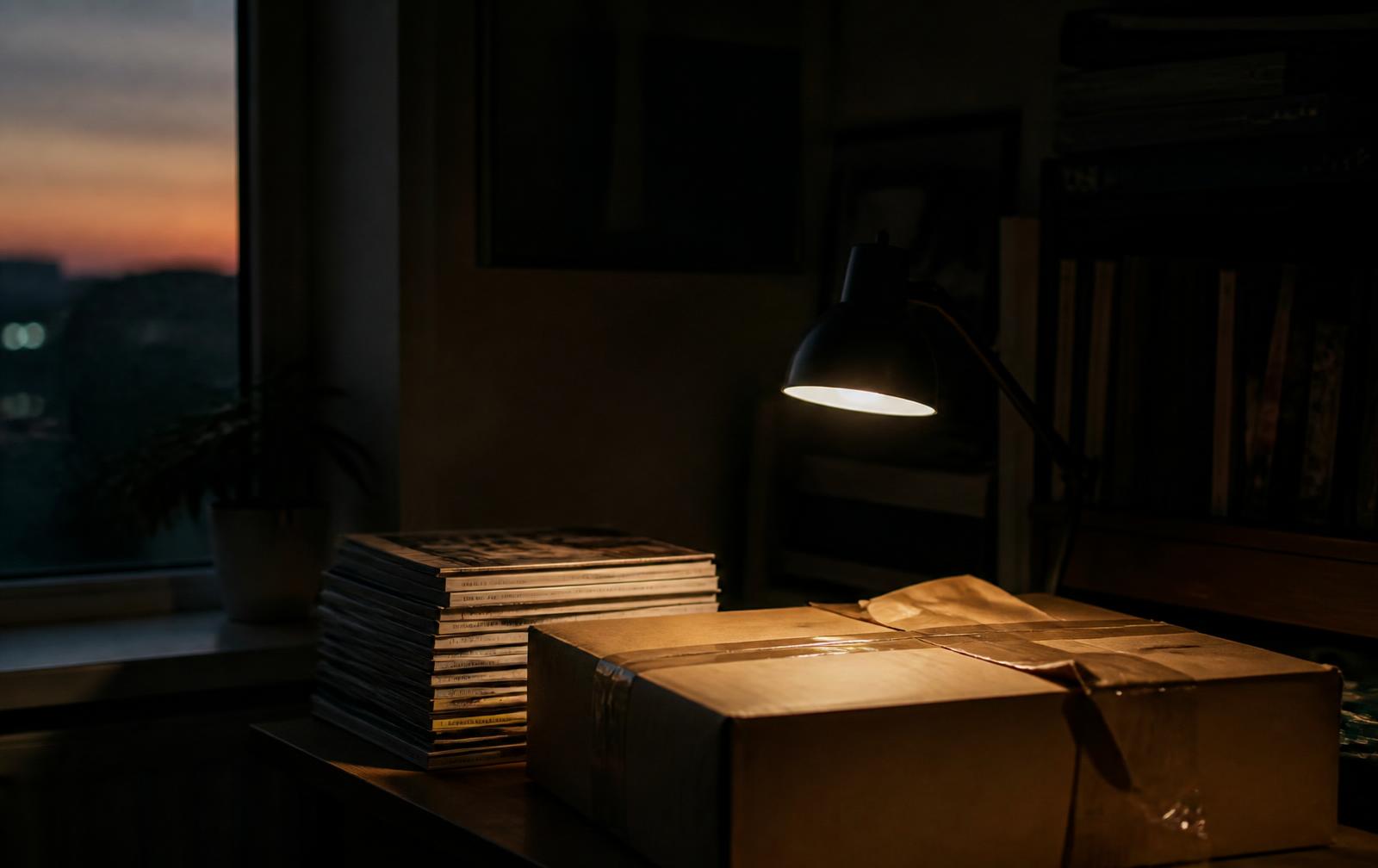 Stack of vintage magazines beside a packed shipping box under a desk lamp at dusk
